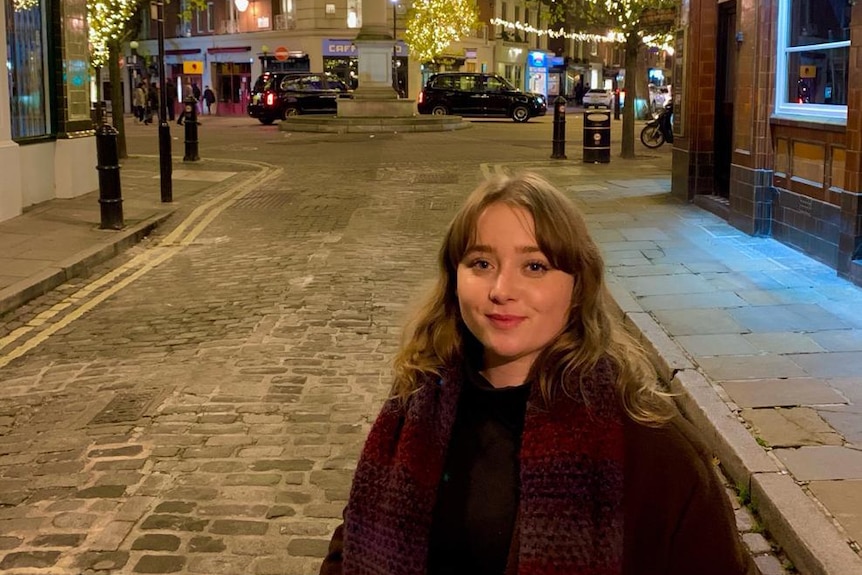 A woman stands in the middle of the street. Behind her is a roundabout with lights decorating the trees.