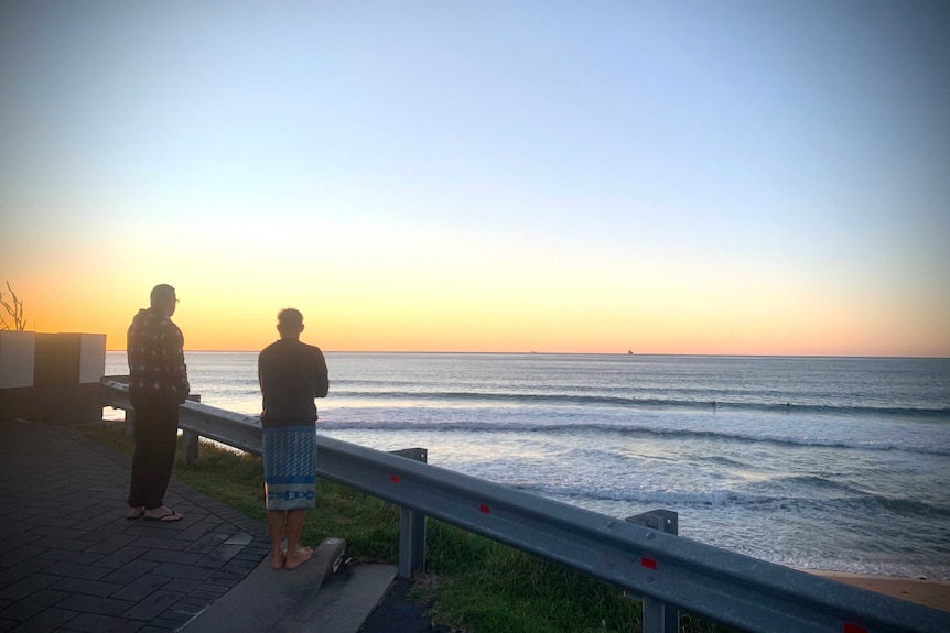 Two surfers stand in a car park overlooking a beach at dawn.