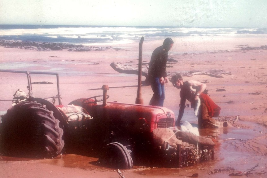 Two people working to get a bogged red tractor out of sand with waves rolling in behind.