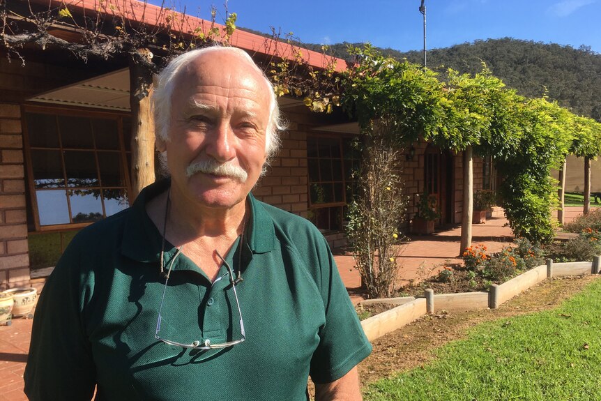 An older man with with snowy hair and a moustache stands in front of a rural property, smiling in the sun.
