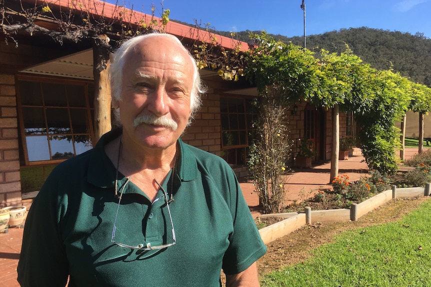 An older man with with snowy hair and a moustache stands in front of a rural property, smiling in the sun.