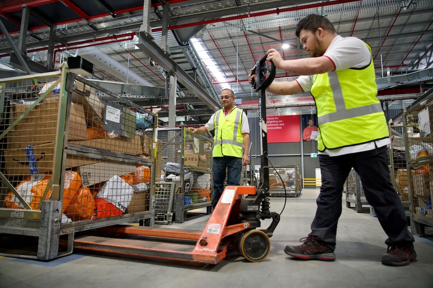 A man using a handheld forklift to lift a pallet of parcels while another man watches on. 
