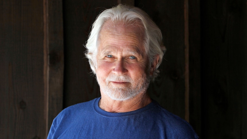 A well-groomed older man with white hair and stubble wearing a blue shirt smiles in front of a dark wooden door.