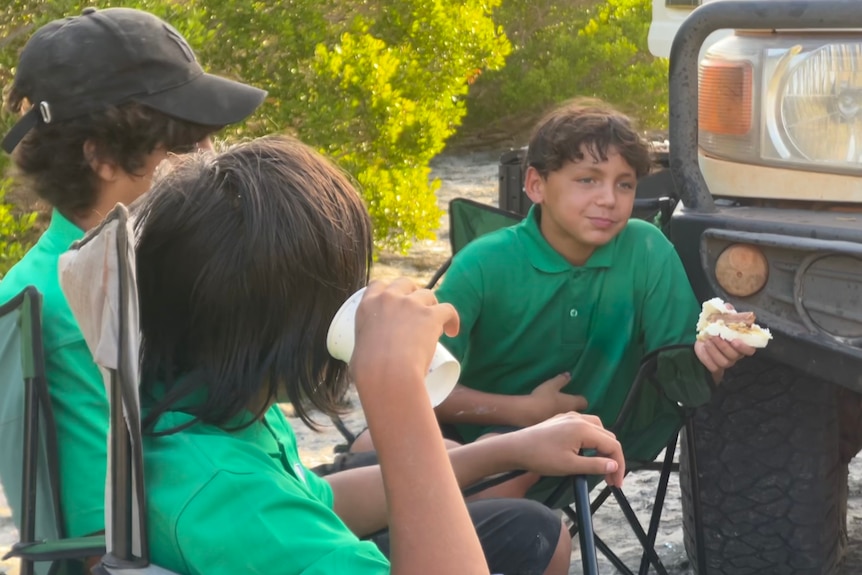 A group of boys sitting around on camping chairs while one eats a sausage.