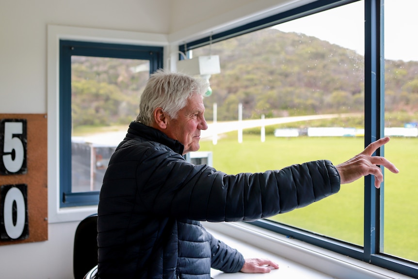 Man with white hair wearing puffer jacket points out window toward football ground