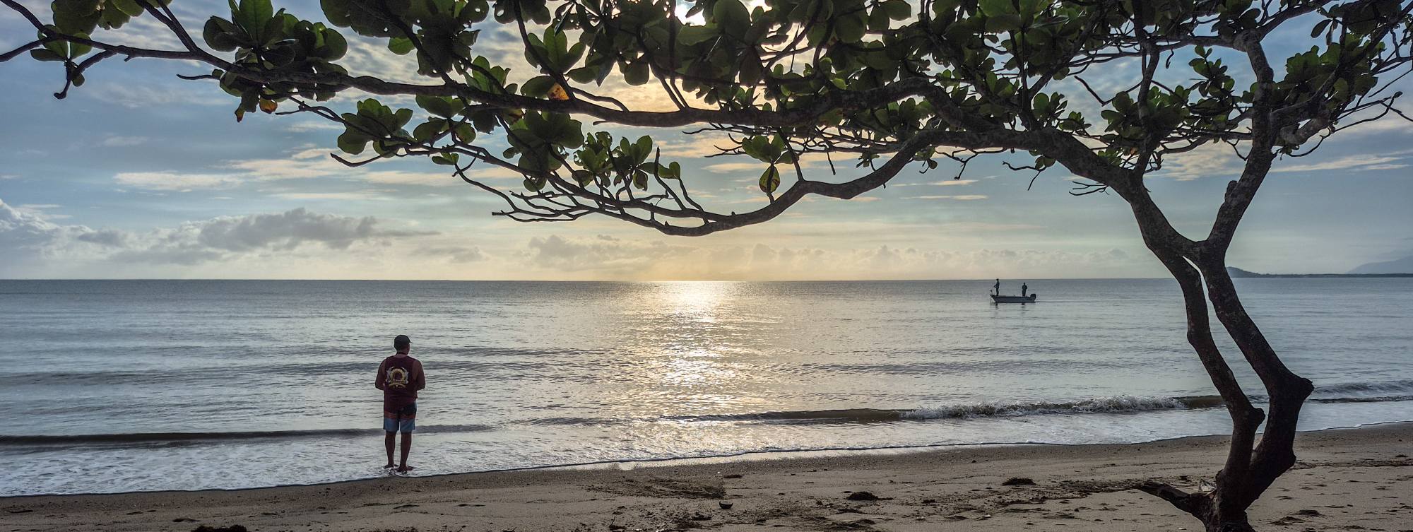 A silhueta de um pescador na praia de manhã cedo. Ao longe estão duas figuras a bordo de um pequeno barco de lata.