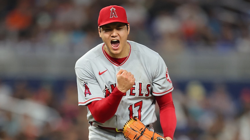 A Japanese man in a grey baseball jersey and red hat pumping his fist in celebration