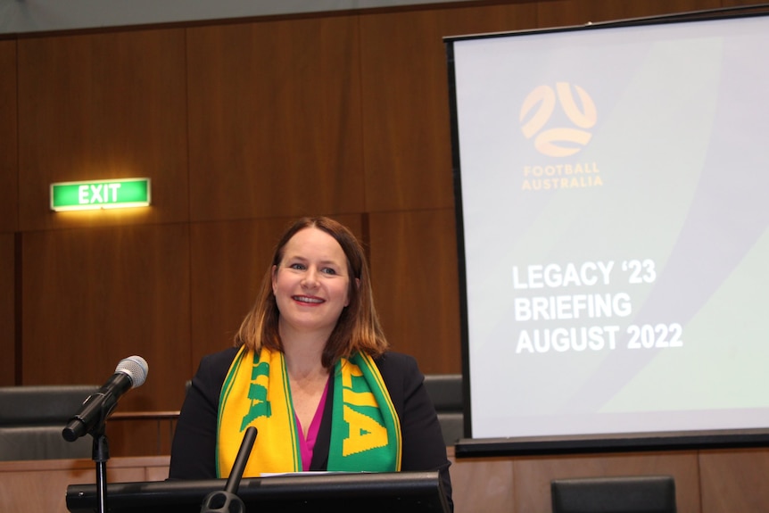 A woman with a Matildas scarf talks in front of a screen.