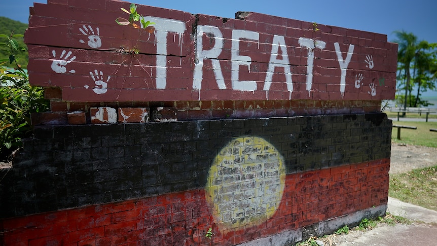 A brick wall says Treaty and has handprints above a painted Aboriginal flag. 