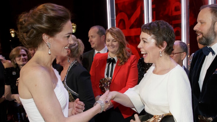 Princess Kae shakes the hand of beaming Olivia Colman while surrounded by people in formal attire.