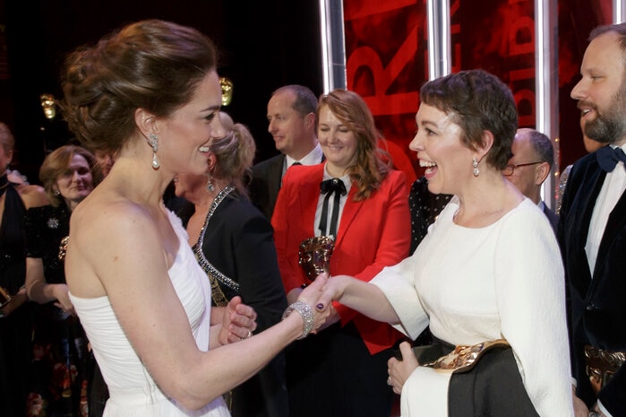 Princess Kae shakes the hand of beaming Olivia Colman while surrounded by people in formal attire.