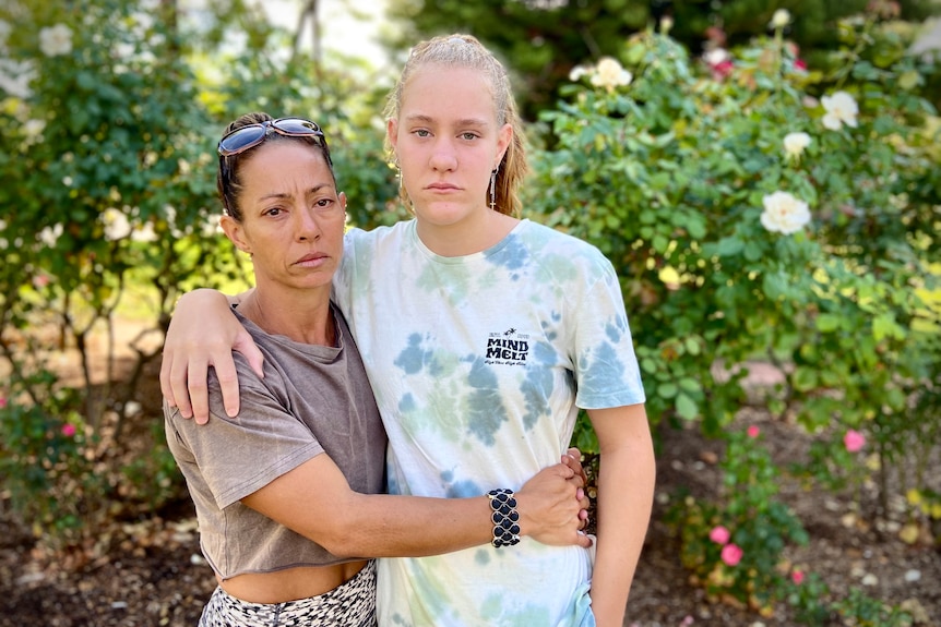 mother wraps her arms around her daughter both looking towards the camera with a solemn look on their faces.