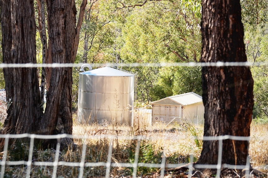 A stainless steel water tank next to a shed, shot from behind a wire fence. 