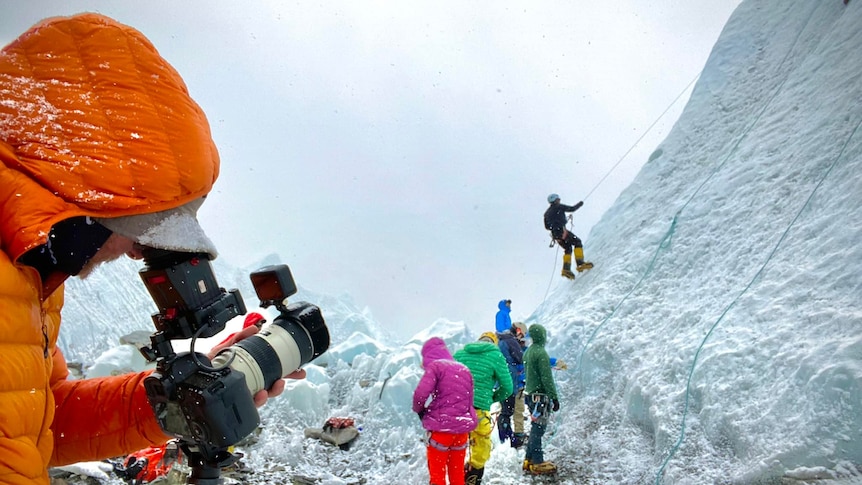 A man films climbers scaling a glacier. 