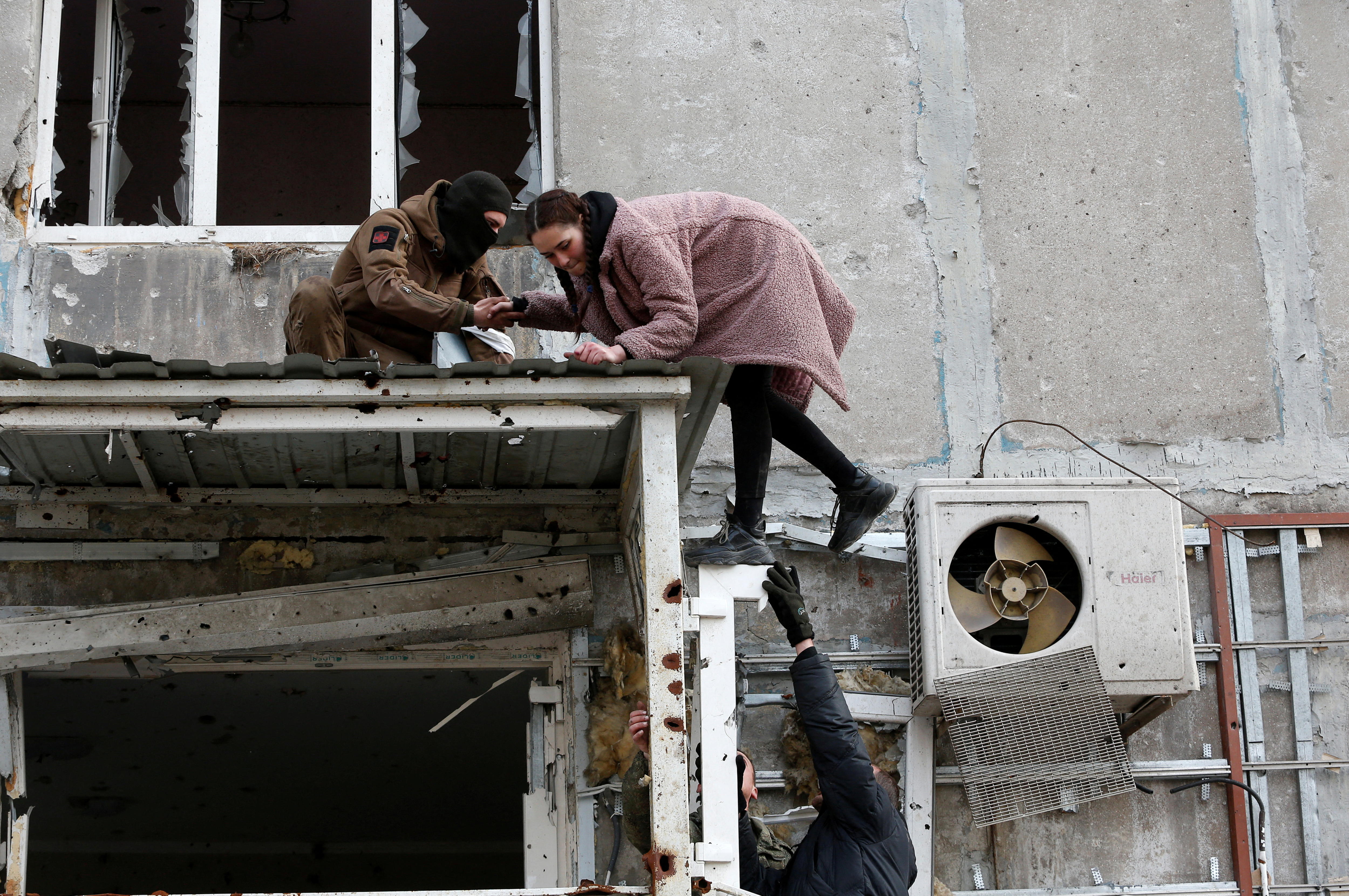 A woman is helped down from an awning near broken windows by two men, one wearing a balaklava