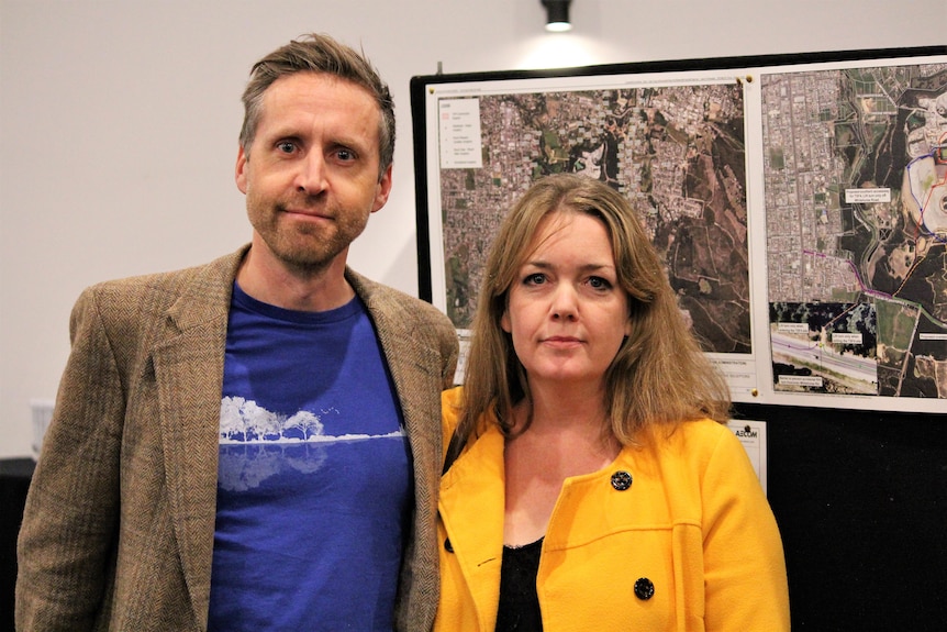 A man and woman stand in front of aerial shots of a gold mine's expansion plans. 