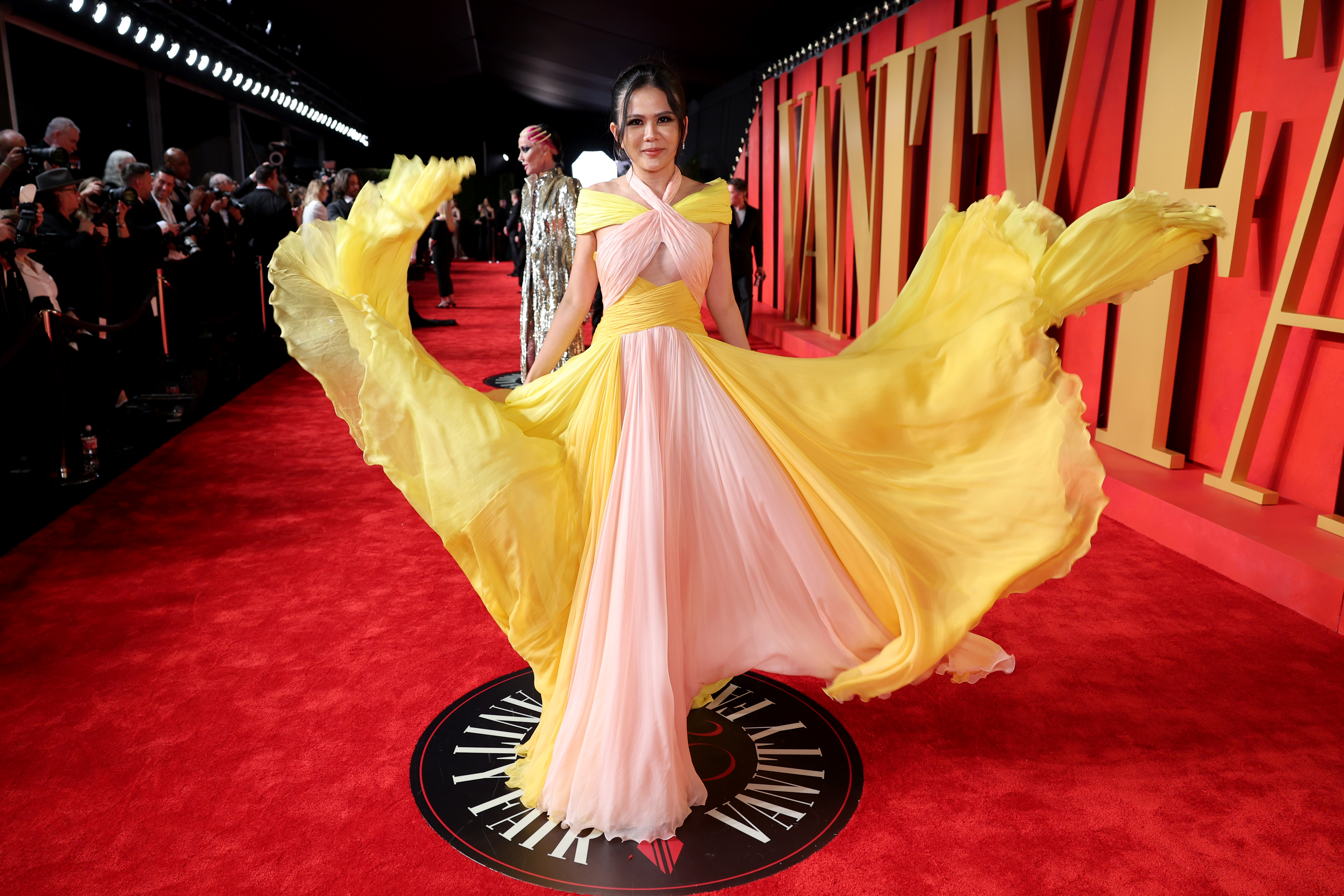 Nichapat Suphap in a yellow and pink flowy dress on a red carpet. 