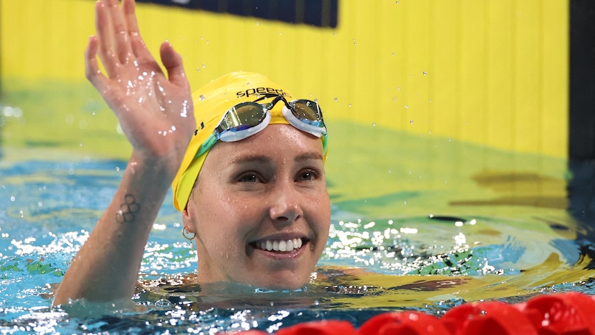 emma mckeon in the pool with her head out of the water waving with her goggles on her forehead