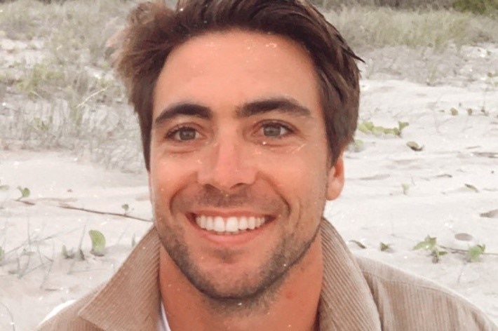 A head shot of a man with brown hair sitting on sand dunes smiling at the camera
