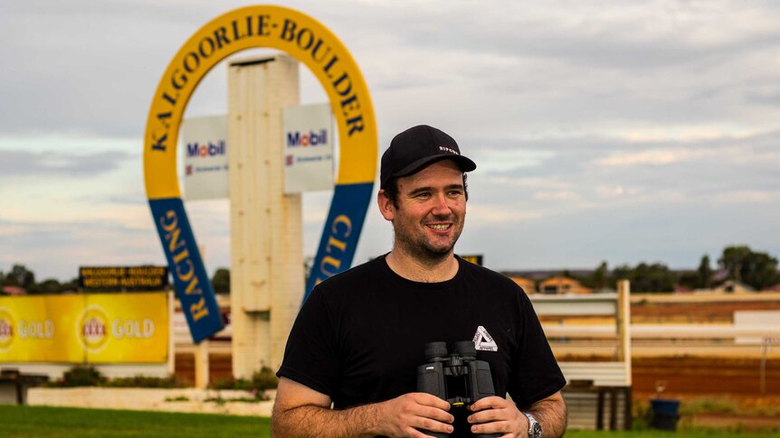 Man holding binoculars on race track with a sign in the background that reads Kalgoorlie-Boulder racing club.