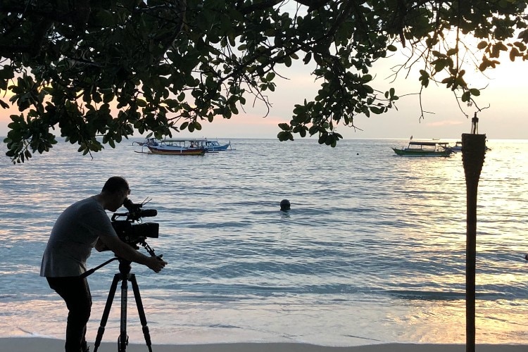 Man operating a camera on a tripod filming a beach at sunset with a person's head above water and two boats in distance.