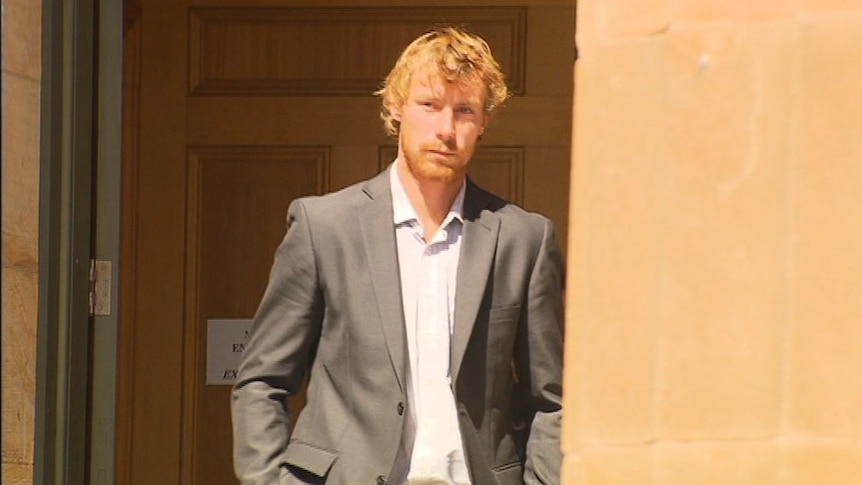 A man with blonde hair and wearing a grey suit walks outside of a courtroom
