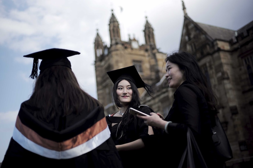 Three women wearing graduation hats and robes outside a sandstone building at the 