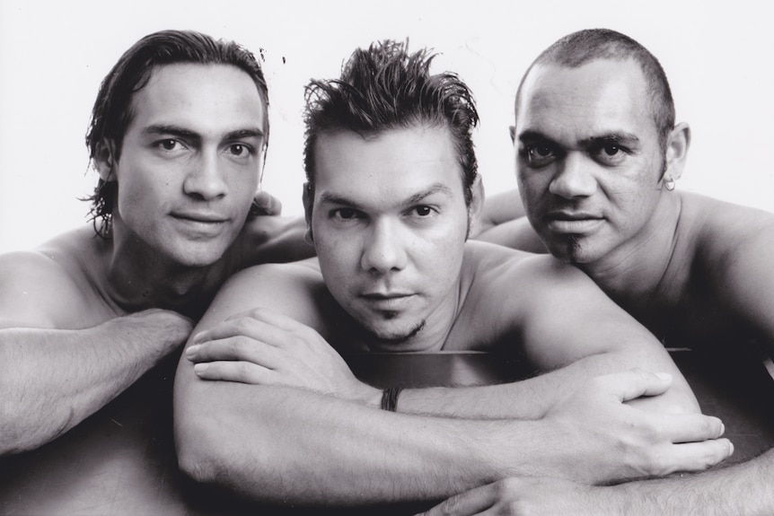 Three young Indigenous men in a black and white 90s photo, they are all shirtless and leaning on each other