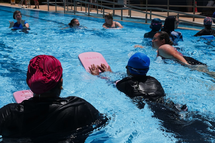 Eight women swim in an outdoor 50 metre pool with an instructor and floatation boards.