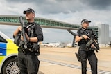 Two police officers standing on an airport runway holding large guns.