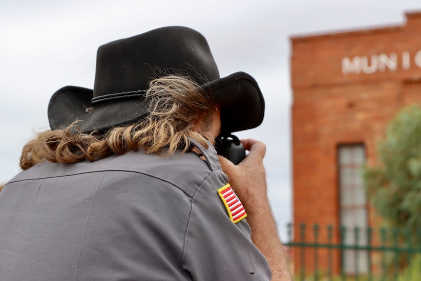 photo from behind of a man with long hair wearing a hat and grey shirt and taking a picture of a brick building 
