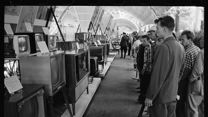 A scan of a black and white negative shows men behind velvet rope looking behind a series of 1950s television sets.