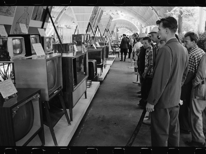 A scan of a black and white negative shows men behind velvet rope looking behind a series of 1950s television sets.