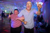 Daz and Gaz give a thumbs up, smiling while dancing in the colourfully lit room.