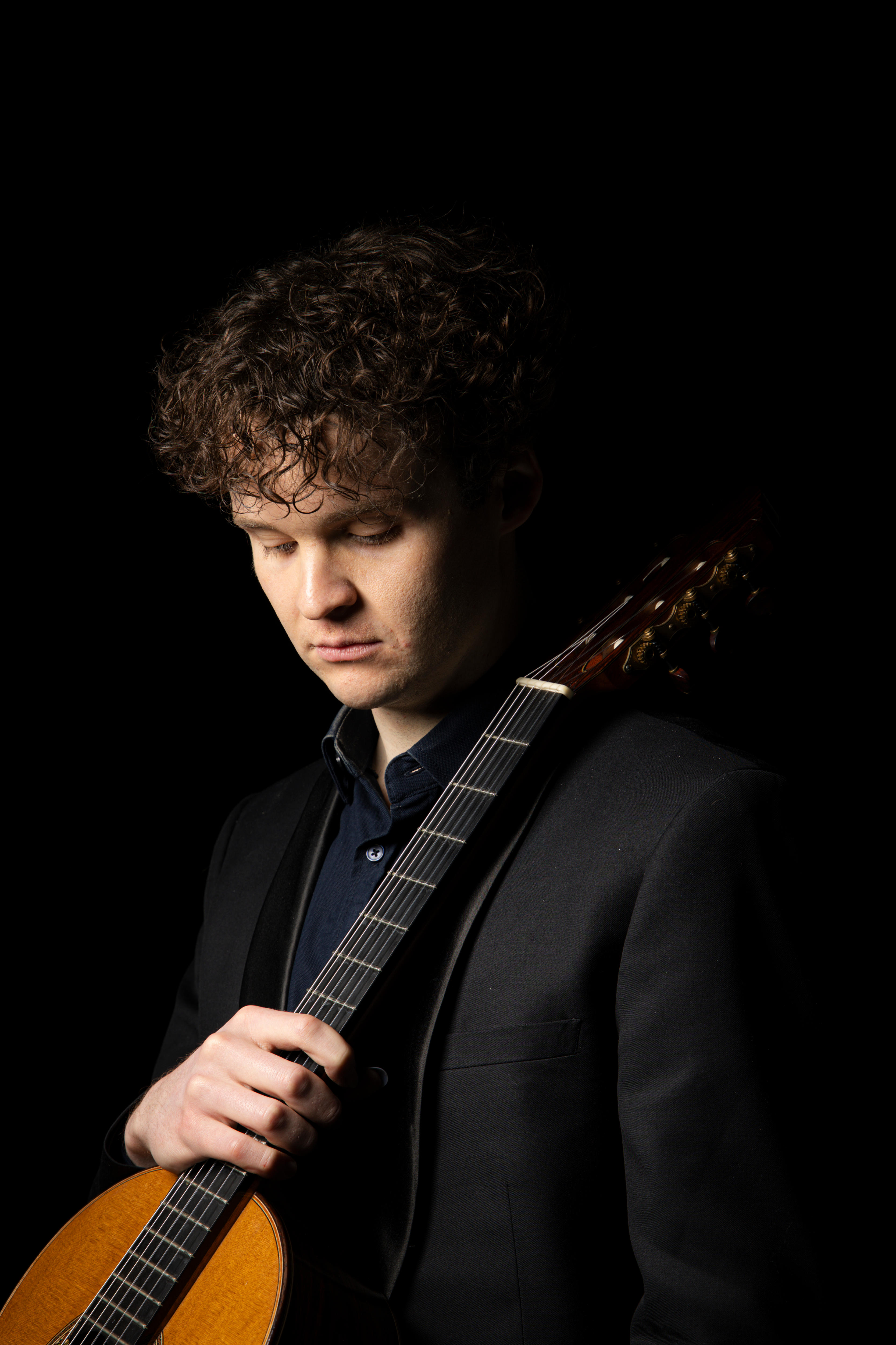 Studio portrait of curly-haired classical guitarist, Connor Whyte holding instrument against shoulder, on a black background.