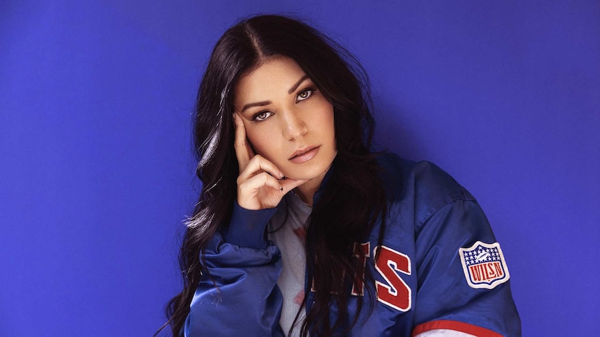 A photo of WILSN with long, dark hair, wearing a baseball jacket, leaning on her fist, not smiling 