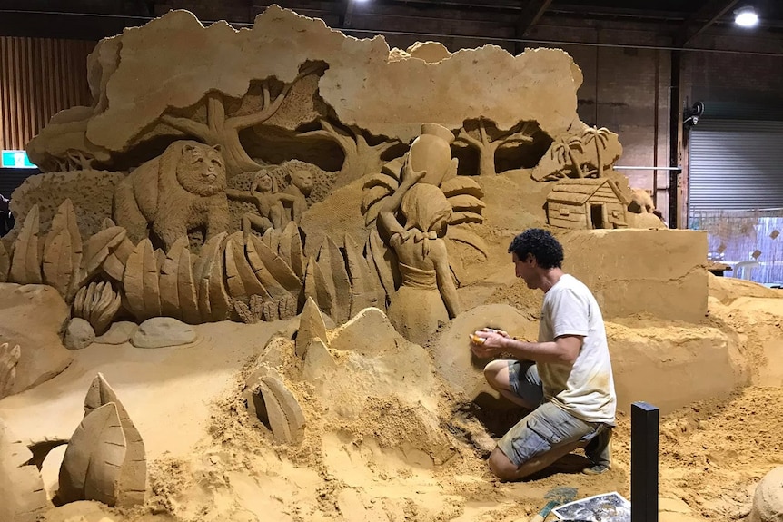 A man in a white shirt and grey shorts with curly dark hair smoothing the surface of a large dark brown jungle sand sculpture.