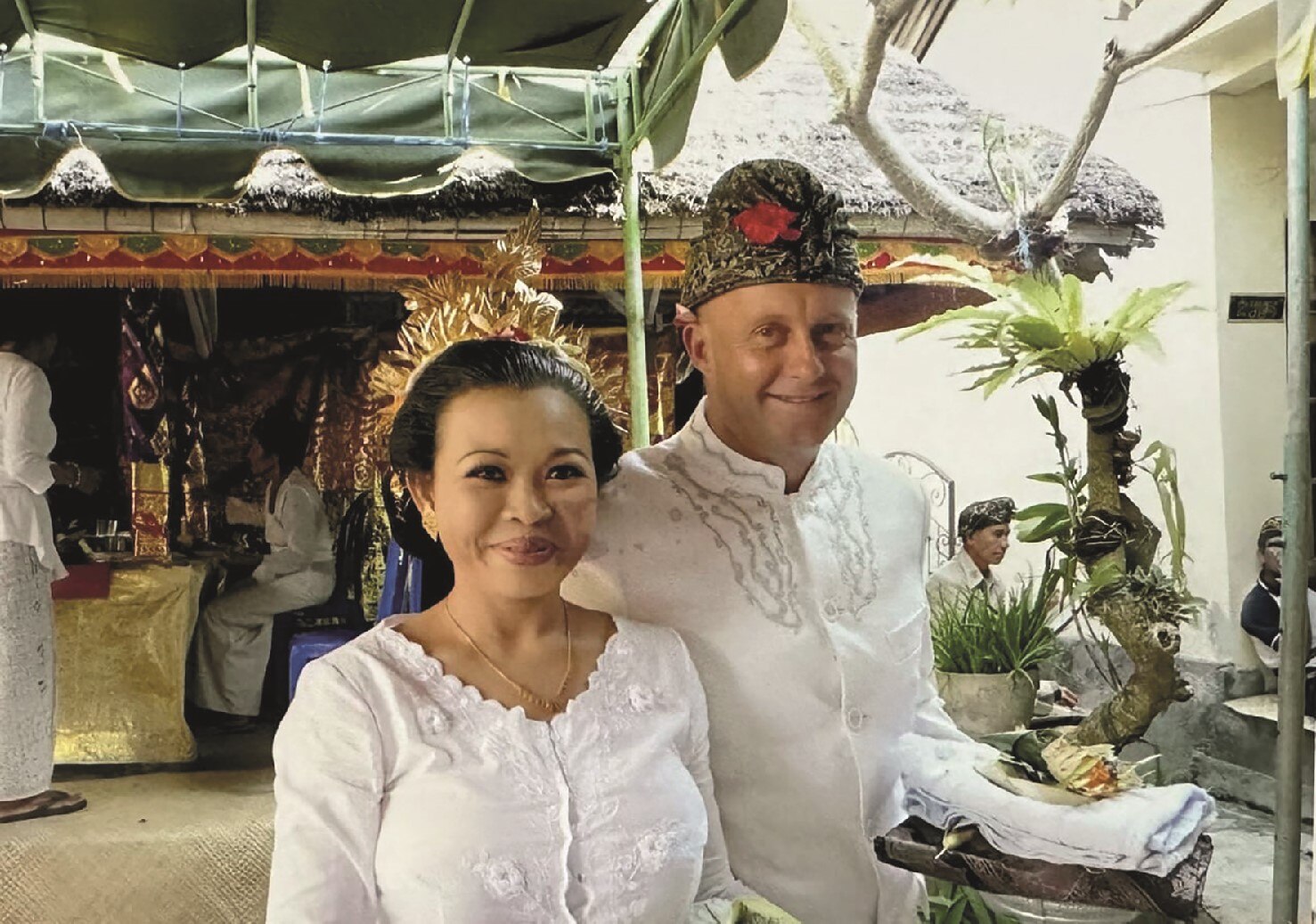 Smiling man, woman in traditional Balinese white dress, embroidered, woman has gold crown, man black print turban with flower.