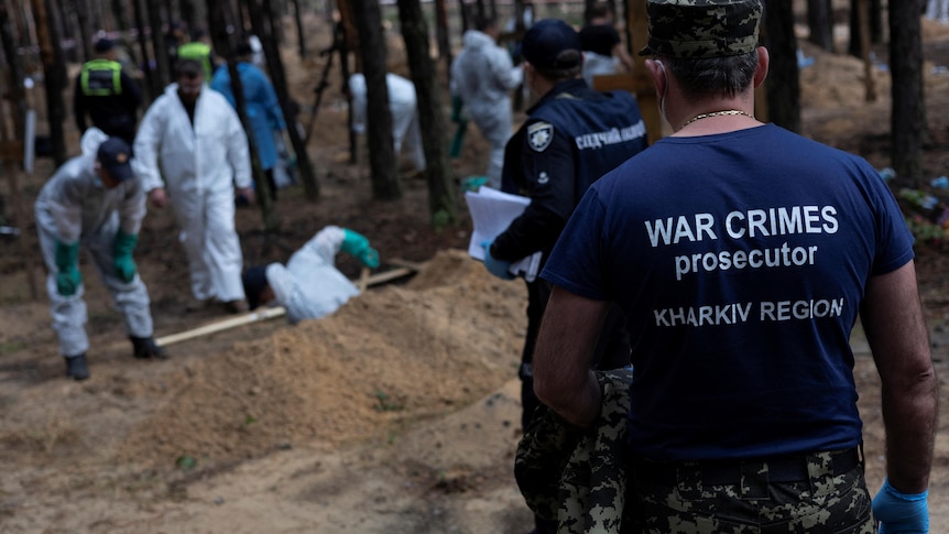 A war crimes prosecutor stands has his job title printed on the back of his shirt, facing a grave site. 