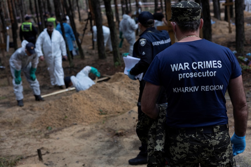 A war crimes prosecutor stands has his job title printed on the back of his shirt, facing a grave site. 