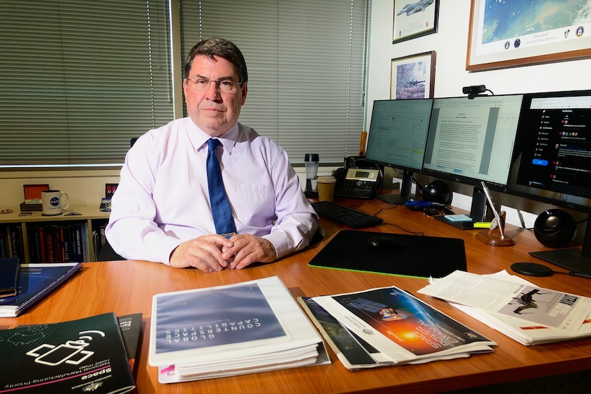 A man in a shirt and tie sitting on a desk with a computer on the right and paperwork in front of him