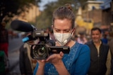 Siobhan Heanue holds a camera while wearing a face mask on a street in India.