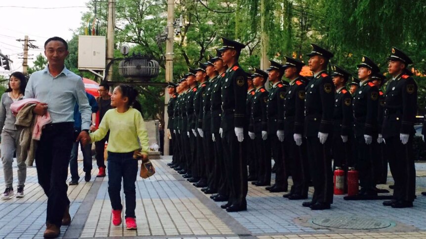 Chinese security forces stand in a Beijing street as pedestrians walk by. They are in full uniform with hands at their sides.