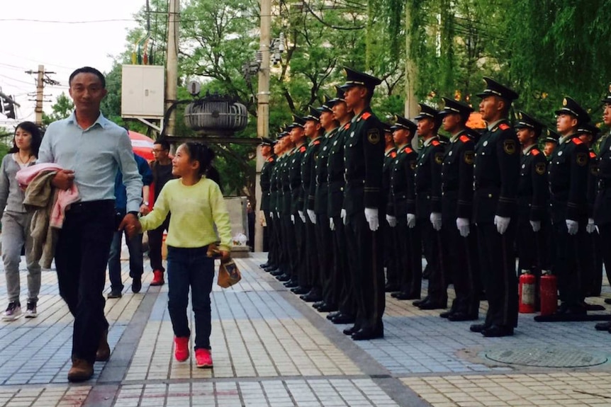 Chinese security forces stand in a Beijing street as pedestrians walk by. They are in full uniform with hands at their sides.