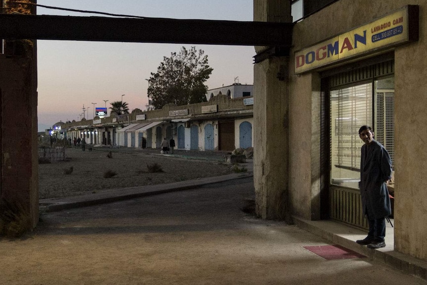 Against the background of a crumbling townscape at dusk the actor stands in front of a rundown shopfront with a Dogman sign.