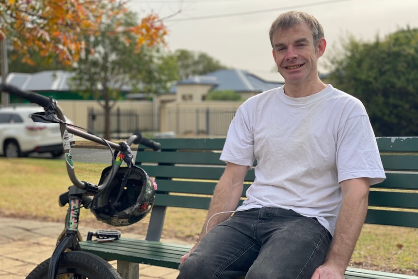 A man sits in a while shirt smiling on a park bench with his bike next to him.