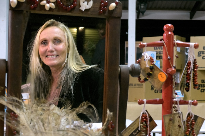 A woman reflected in a mirror, standing behind the counter at a market stall, with bracelets on a rack beside her.