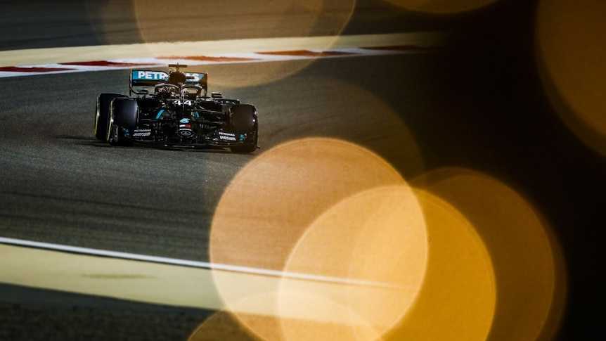 An F1 driver rounds a corner in his car with lights twinkling in the foreground.