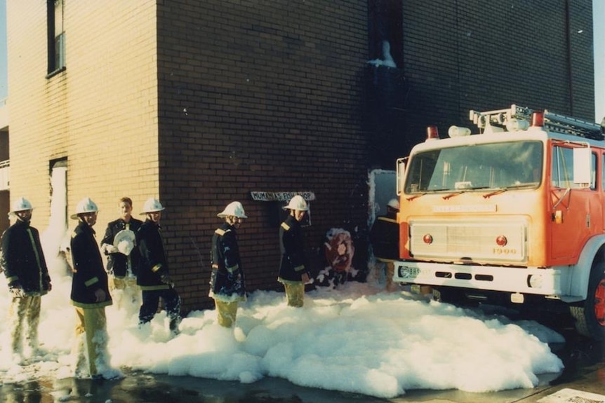 A bunch of firefighters stand around in foam up to their knees near a fire truck.