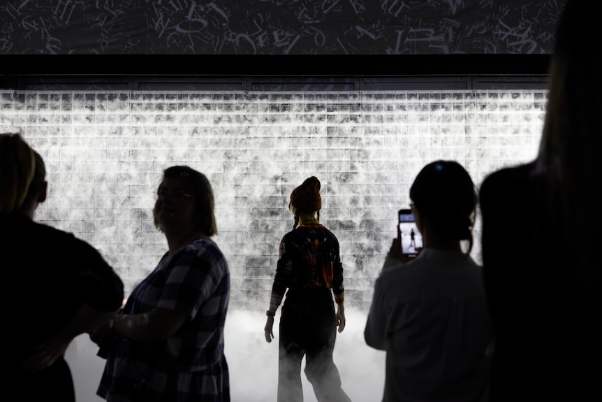 Five people are silhouetted by a large backlit grid with billowing white water vapour flowing out of it.